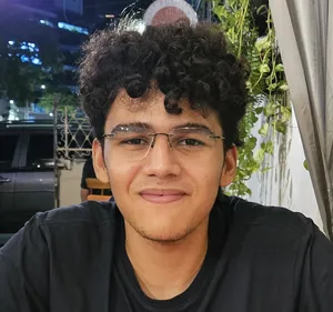 A self-portrait of a young man with curly dark hair and glasses,
        smiling faintly and looking directly at the camera. He is wearing a
        dark t-shirt and is seated outdoors at night, with blurred lights and
        structures in the background.